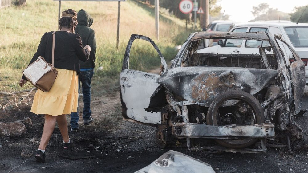 Pessoas indo para o trabalho passam por um carro queimado depois que multidões enfurecidas o incendiaram quando o presidente Cyril Ramaphosa visitou a província de Kwazulu-Natal, onde a violência continuou por 6 dias. (Photo by RAJESH JANTILAL / AFP)