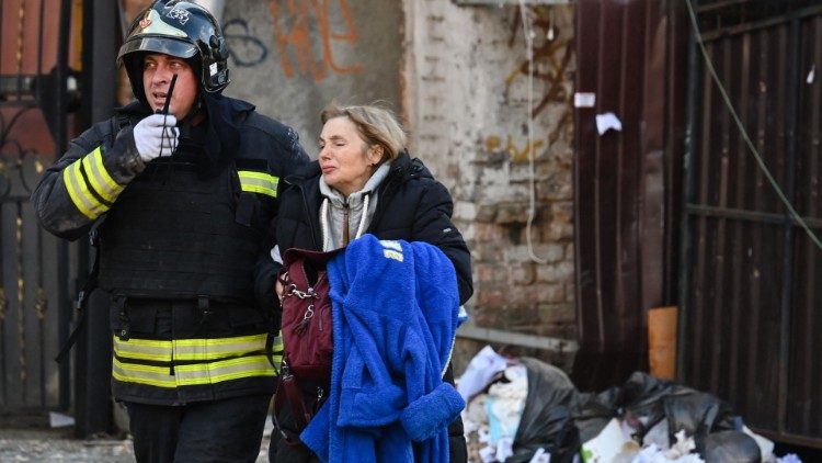 Um socorrista ajuda uma moradora local enquanto ela caminha do lado de fora de um prédio de escritórios de vários andares parcialmente destruído ao sair após vários ataques russos atingirem a capital ucraniana de Kyiv em 10 de outubro de 2022, em meio à invasão da Ucrânia pela Rússia. (Photo by Sergei SUPINSKY / AFP)